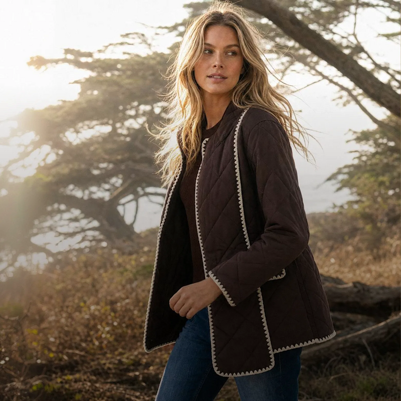 A woman wearing a dark brown quilted jacket with cream-colored trim, standing outdoors in a sunlit forest setting.