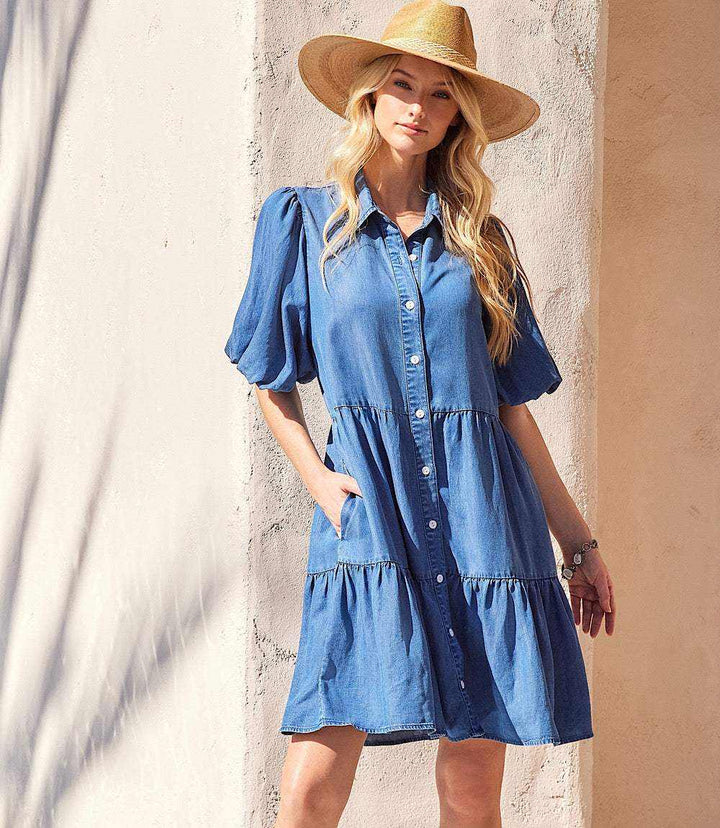 Woman wearing blue puff sleeve tiered shirtdress with front buttons and side pockets, accessorized with a straw hat.