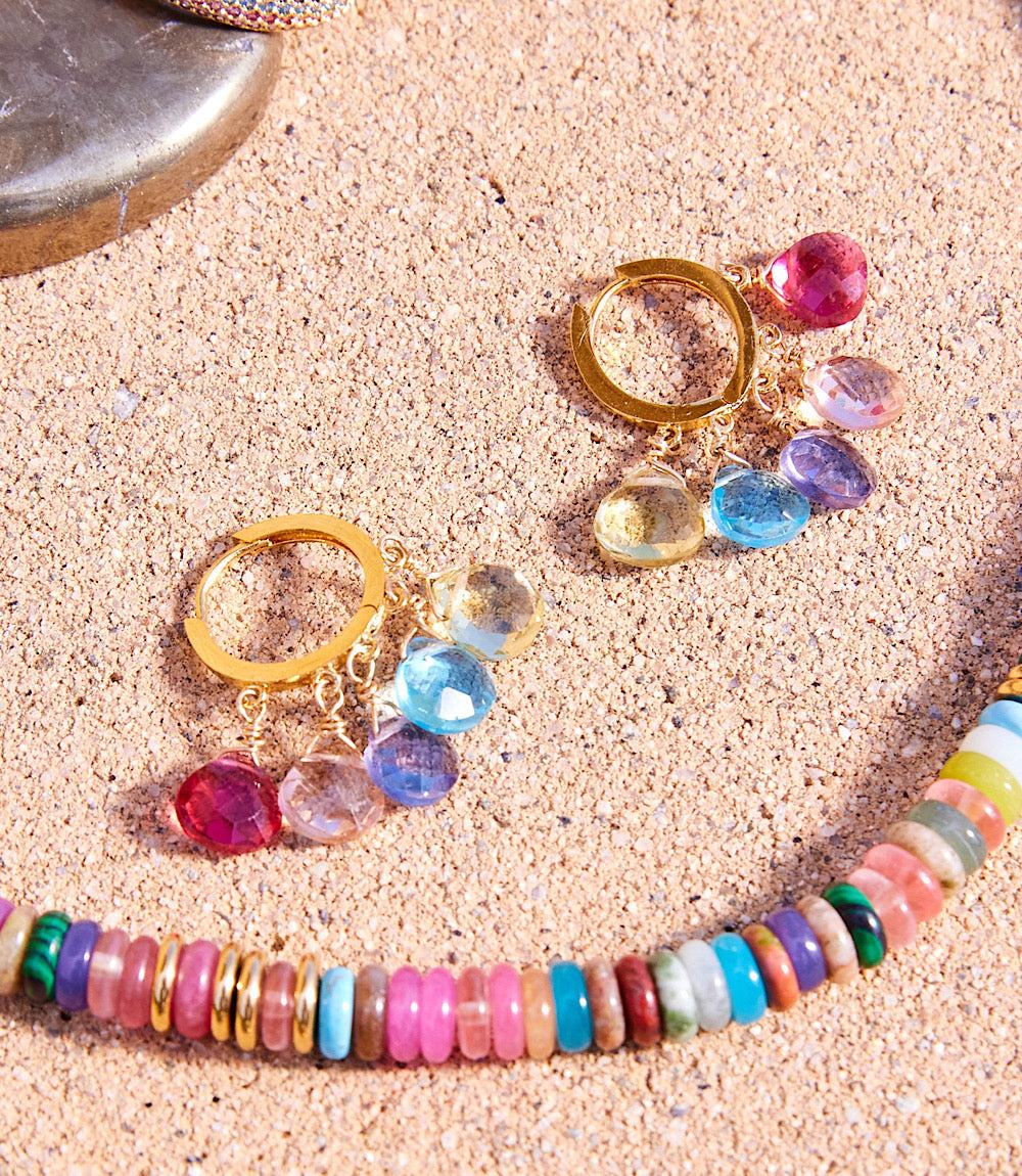 Gold gemstone huggie earrings with rose quartz, aquamarine, pearl, and labradorite charms on sandy background with colorful beaded necklace