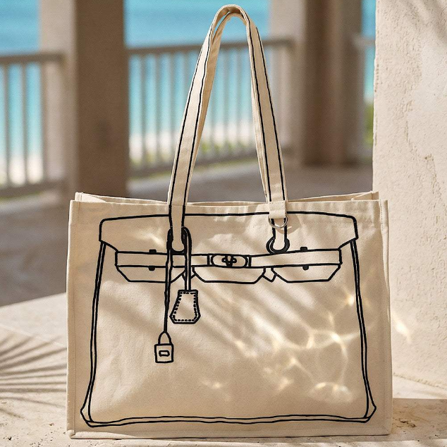 Beige tote bag with black outline of a handbag design on a sandy surface.