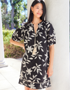 Woman wearing a black dress with white floral pattern standing against a white wall.