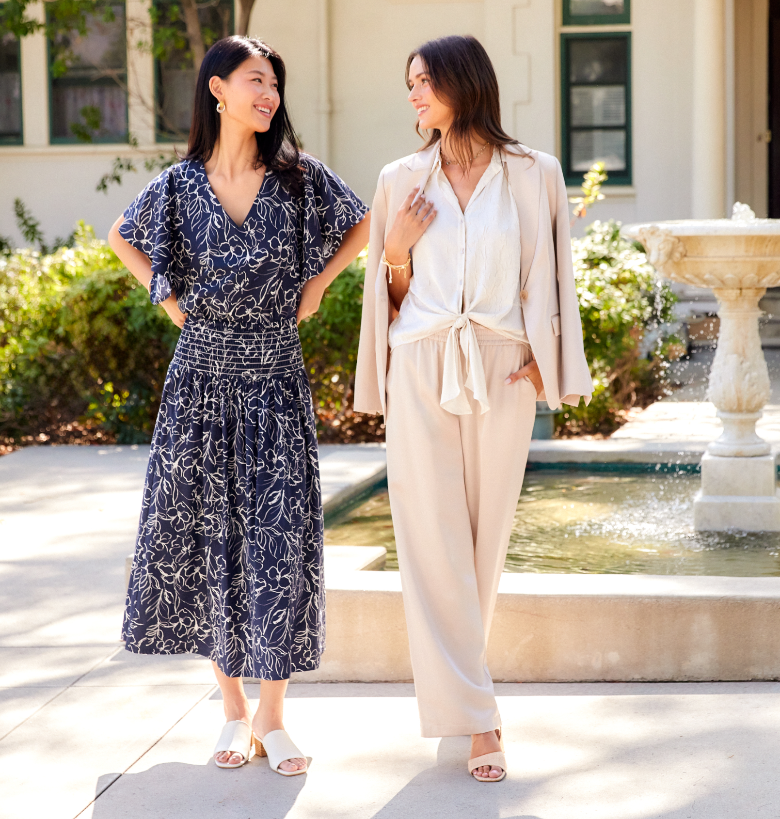 Two women standing outdoors by a fountain, one in a navy patterned dress and the other in a beige outfit.