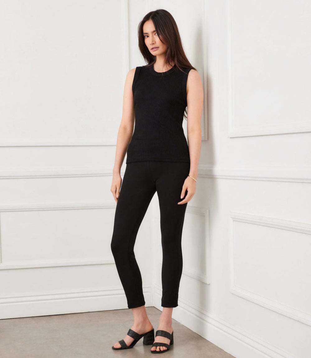 Woman wearing black skinny ankle-length Piper Pants with sleeveless black top and black sandals in minimalist room