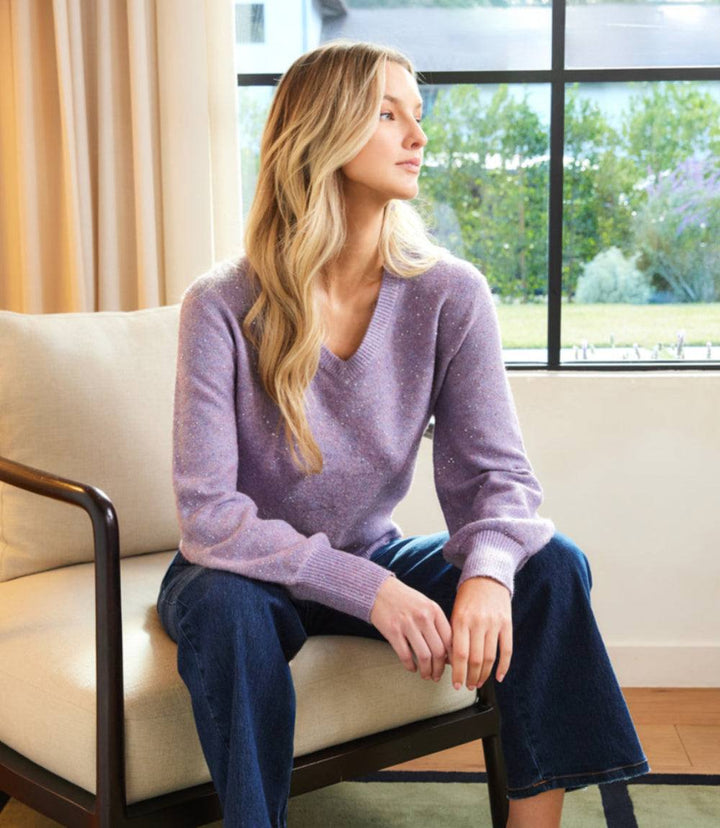 Woman wearing lavender sequin V-neck sweater with ribbed edges and drop shoulder sleeves sitting indoors