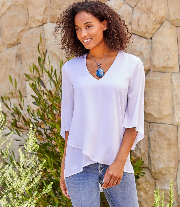 Woman wearing a white silky crepe Bracelet Sleeve Asymmetric Top with V-neck, flare sleeves, and layered asymmetrical hem.