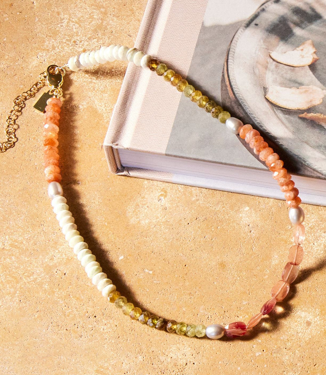 Beaded gemstone necklace with lemon chrysoprase, green garnet, sunstone, grey pearl, andalusite, and 14k gold vermeil on a beige surface next to a book.