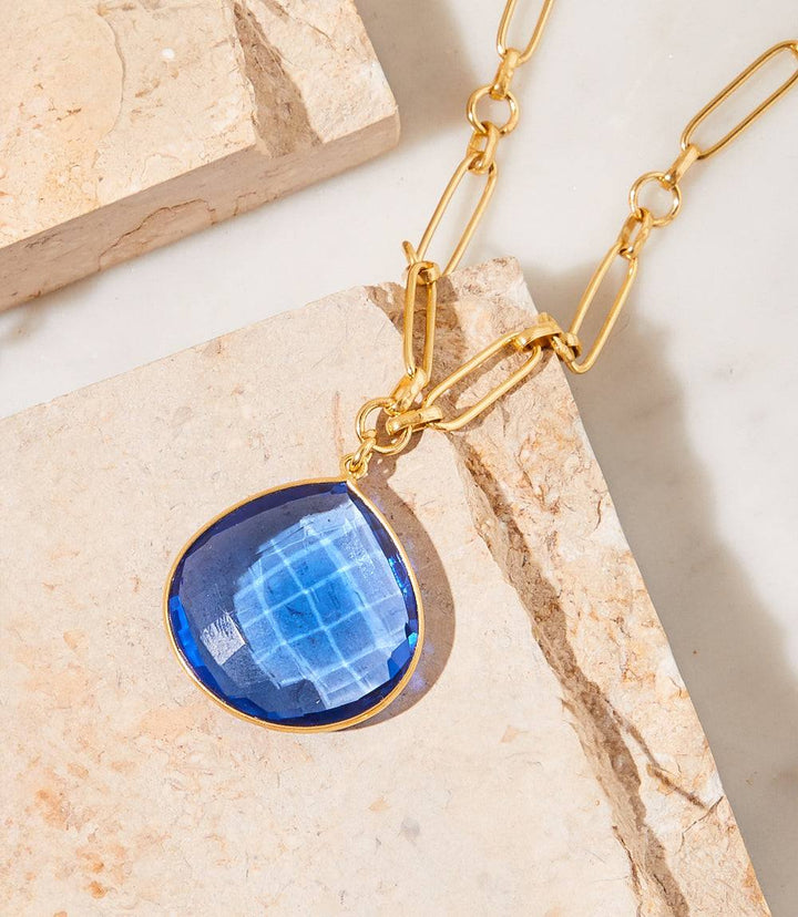 Blue tanzanite pendant necklace with gold chain on beige stone background