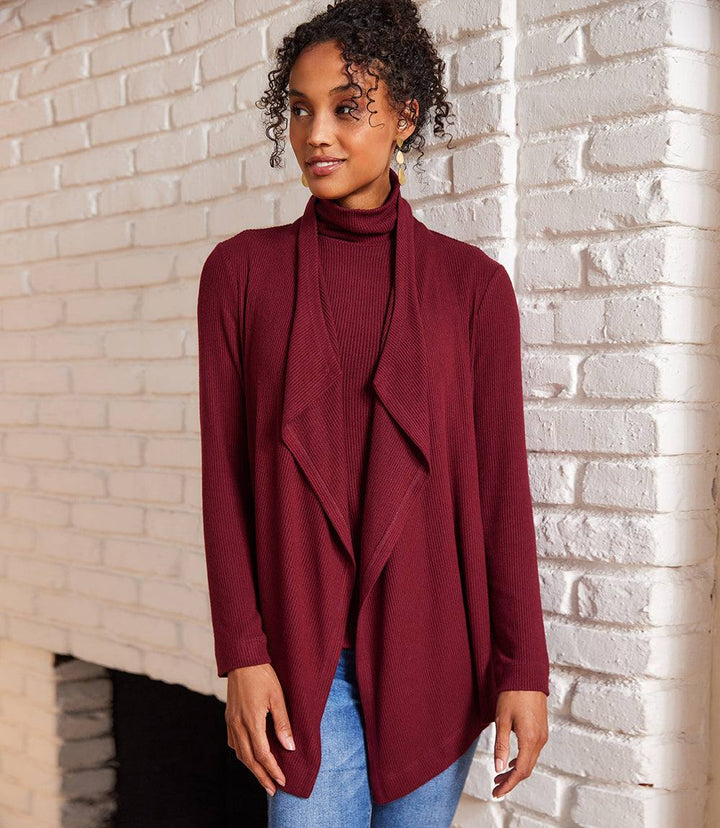 Woman wearing a maroon long sleeve ribbed drape front cardigan with open front and relaxed fit standing against white brick wall