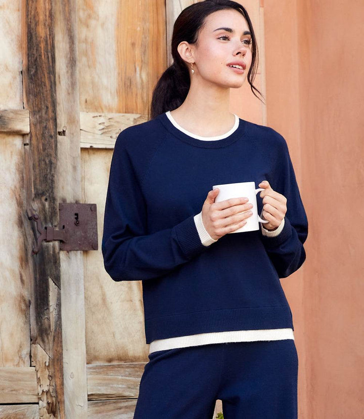 Woman wearing navy contrast crewneck sweater with raglan sleeves holding a white mug standing by wooden door