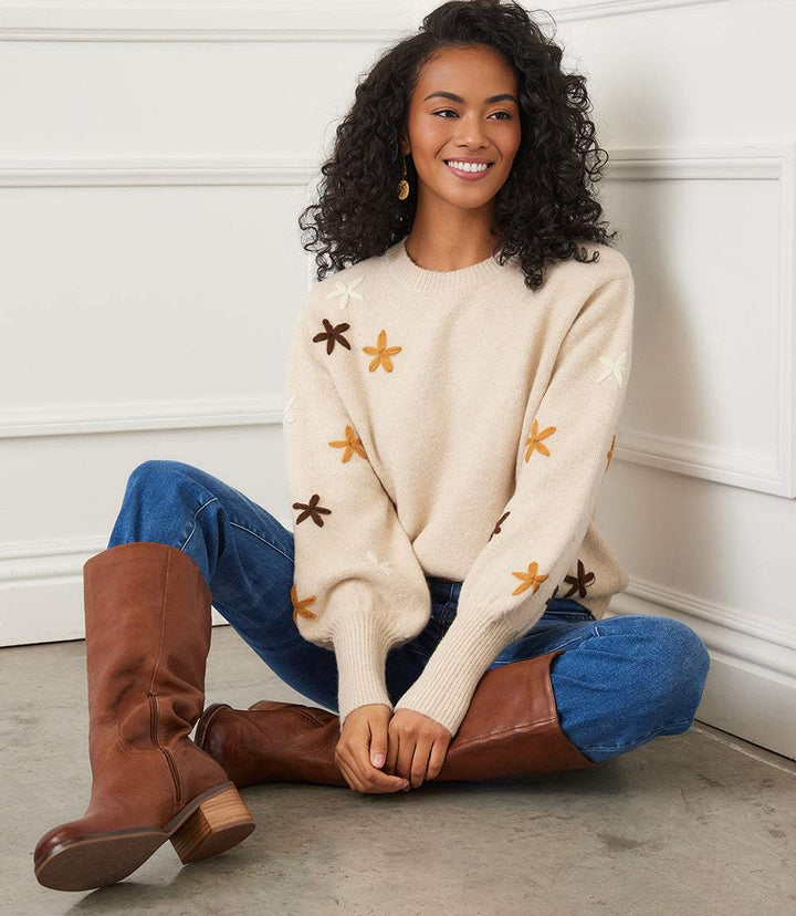 Woman wearing beige embroidered flower sweater with extended sleeves and blue jeans sitting on floor