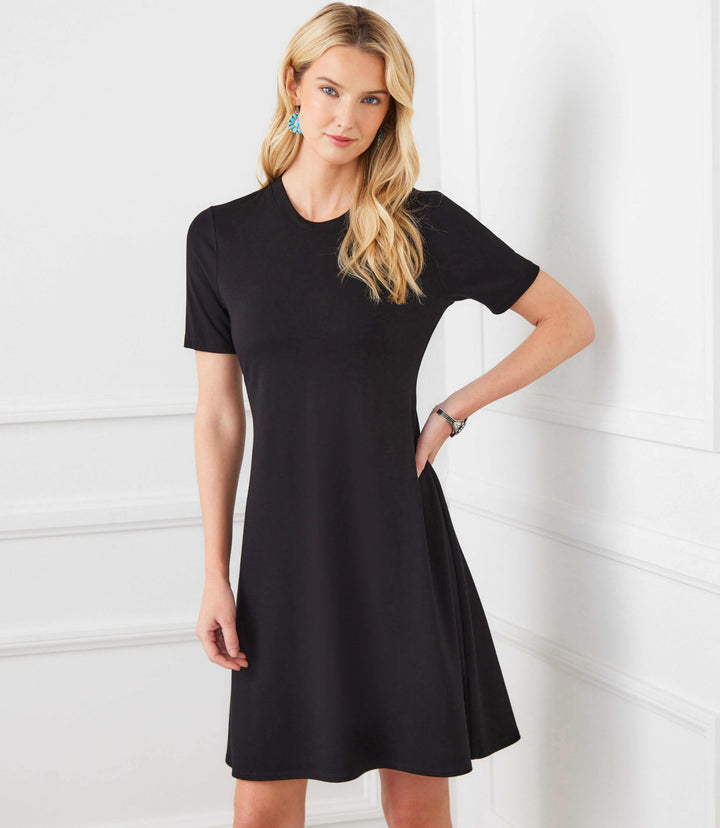 Woman wearing a black short sleeve A-line dress with crew neckline, styled simply in a bright room
