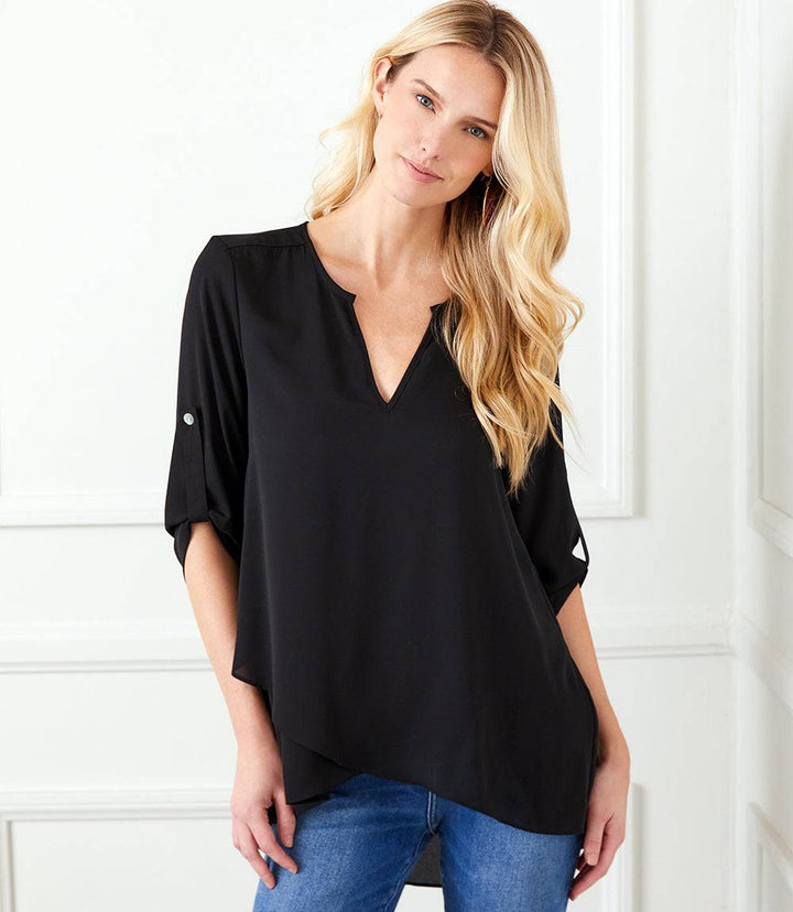 Woman wearing a black asymmetric hem wrap top with split neckline and button tab sleeves paired with blue jeans