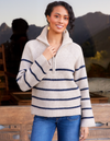 A woman wearing a ribbed knit half-zip sweater with horizontal navy blue stripes on a cream base, paired with blue jeans, set against a rustic outdoor background with mountains and wooden furniture.