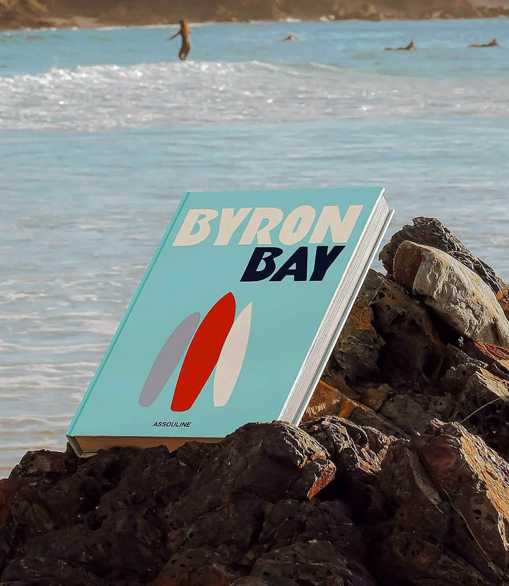 Byron Bay travel book hardcover on rocks by ocean with surfers in background