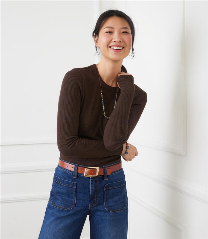 Woman wearing a fitted long sleeve ribbed crewneck top in dark brown paired with blue jeans and a brown belt.