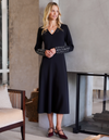 A woman models a long-sleeve black midi dress with intricate silver beaded detailing on the cuffs, styled with burgundy heels in a modern indoor setting.