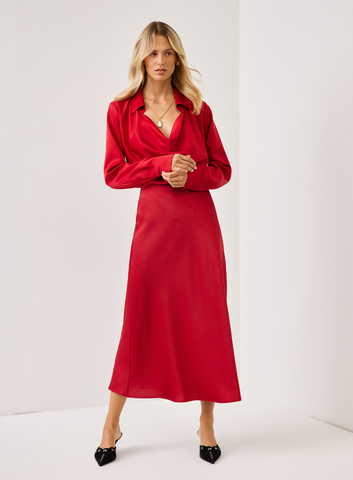 A woman modeling a long-sleeved red midi dress with a collared V-neck, paired with black pointed-toe heels, in a bright studio setting.