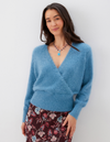 A woman wearing a fuzzy light blue long-sleeve V-neck wrap sweater and a dark floral skirt, standing against a plain light gray wall.