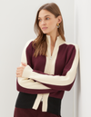 A woman wearing a color-blocked knit zip-up sweater with burgundy and cream panels, standing with arms crossed against a plain light gray wall.