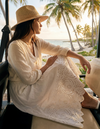 Woman in a white dress and straw hat sitting by a beach with palm trees.