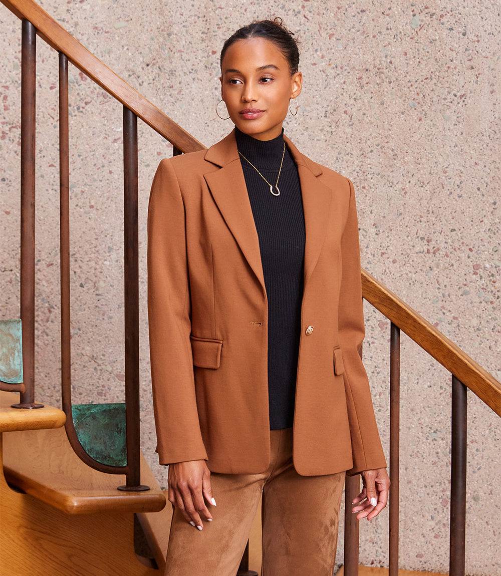Woman wearing a fitted brown Ponte knit blazer with gold buttons, black turtleneck, and brown pants standing by wooden stairs.