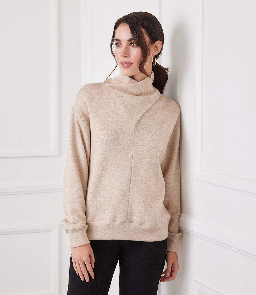 Woman wearing beige soft sweater knit faux wrap turtleneck with asymmetrical neck and dolman sleeves.