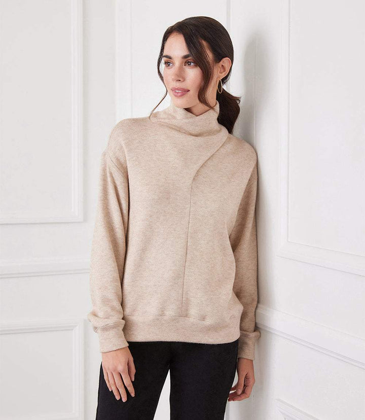 Woman wearing beige soft sweater knit faux wrap turtleneck with asymmetrical neck and dolman sleeves.