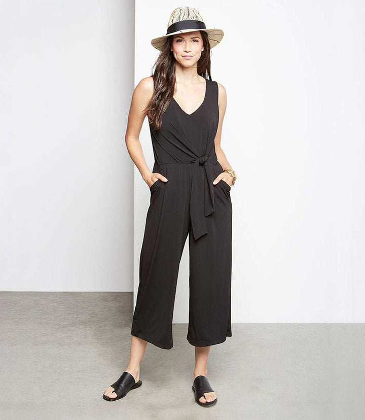 Woman wearing petite size black tie-front travel jumpsuit with cropped legs, side pockets, and sleeveless design, paired with a hat and black slip-on sandals.