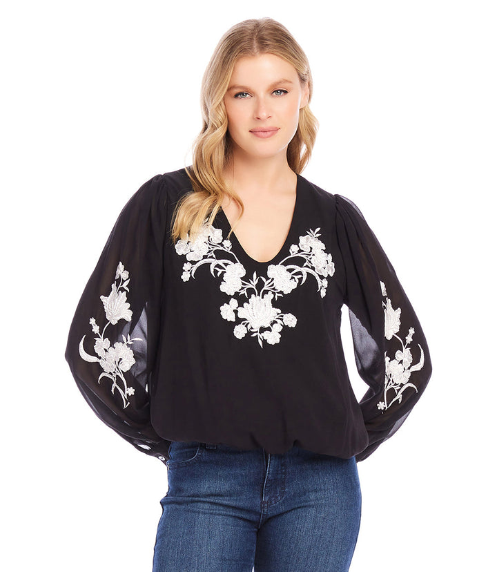 Woman wearing black embellished blouson top with white floral embroidery and sheer sleeves.