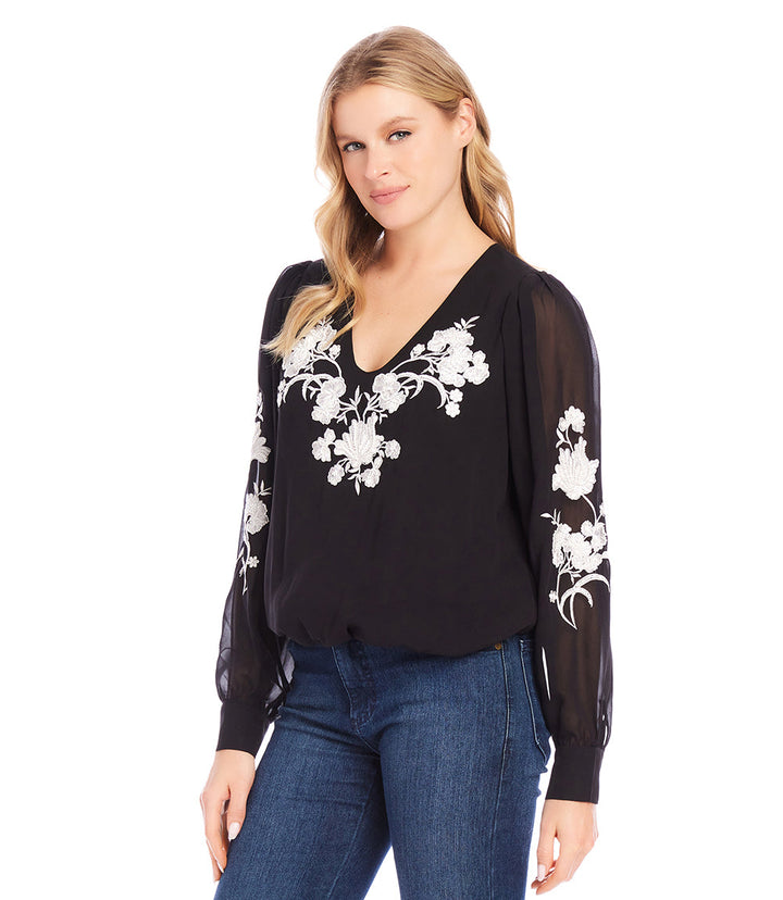Black embellished blouson top with white floral embroidery and sheer sleeves worn with jeans