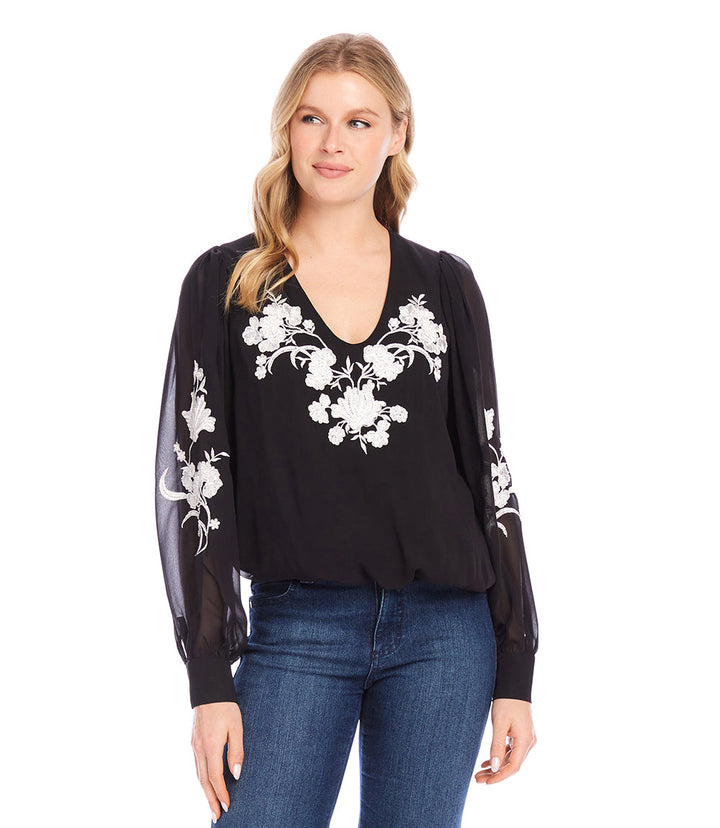 Woman wearing black embellished blouson top with white floral embroidery and sheer sleeves paired with blue jeans