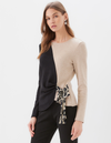 A woman modeling a color-block knit sweater with a split design: one half is black and the other half is beige. The beige side has a tie detail at the waist with a black and beige patterned fabric.