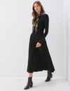 A woman models a long-sleeve black ribbed midi dress with a high neckline, styled with a black, gold, and blue patterned scarf and black ankle boots against a clean white studio background.