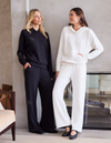 Two women modeling matching lounge sets in a modern indoor setting. One wears a black hoodie and wide-leg pants with small rhinestone embellishments, and the other wears a white matching hoodie and wide-leg pants.