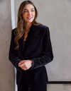 A woman wearing a black velvet blazer over a black top, standing against a textured gray concrete wall.