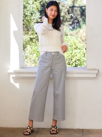 Woman wearing a white sweater and striped pants standing by a window with greenery outside.