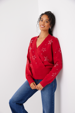 A woman wearing a bright red knit wrap sweater with embroidered white stars, paired with blue jeans, leaning against a white wall.