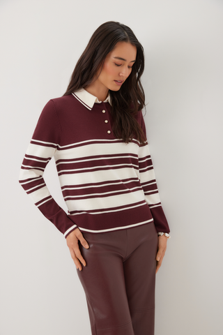 A woman wearing a burgundy and cream striped knit polo sweater with matching brown pants, standing against a plain light gray studio background.