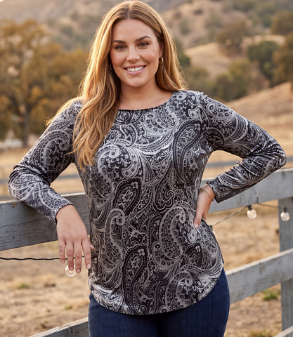 A woman wearing a long-sleeve scoop-neck top with a black and gray paisley pattern, standing outdoors in front of a wooden fence and dry hills.