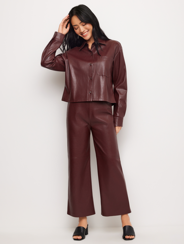 A woman modeling a matching two-piece faux leather outfit in a deep burgundy color, consisting of a cropped button-up shirt and wide-leg pants, paired with black slide sandals against a plain light gray background.