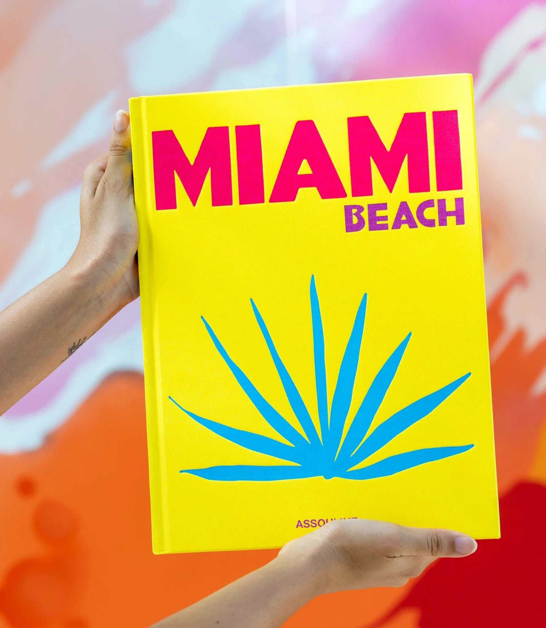 Bright yellow hardcover coffee table book titled Miami Beach with blue palm leaf graphic being held by hands against colorful background