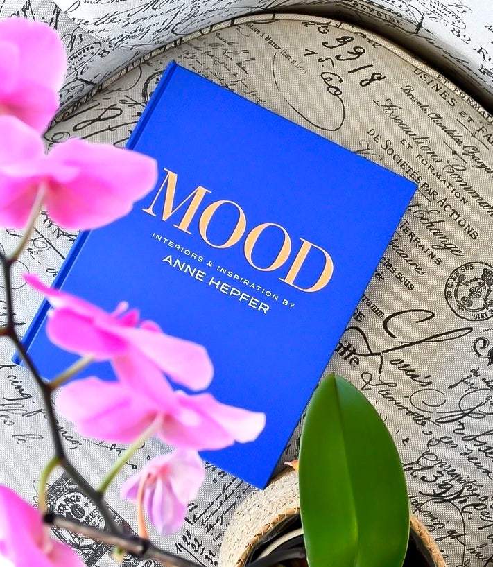 Blue hardcover book "Mood: Interiors and Inspiration" by Anne Hepfer on a patterned fabric chair with pink orchid flowers in the foreground.