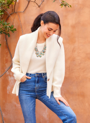 Woman wearing a white sweater, blue jeans, and a colorful necklace against an orange wall.