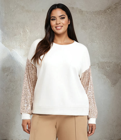 Woman wearing a white sweater with glittery sleeves against a textured wall.