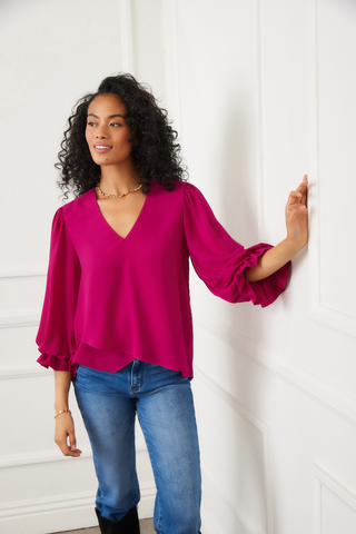 Woman wearing a bright pink blouse and blue jeans against a white wall.