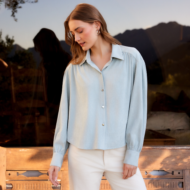 A woman wearing a light blue long-sleeve button-up blouse with gathered shoulders and white pants, standing outdoors in front of a wooden railing and a scenic background.