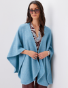 A woman wearing a light blue open-front poncho over a patterned blouse, standing against a plain light gray wall.