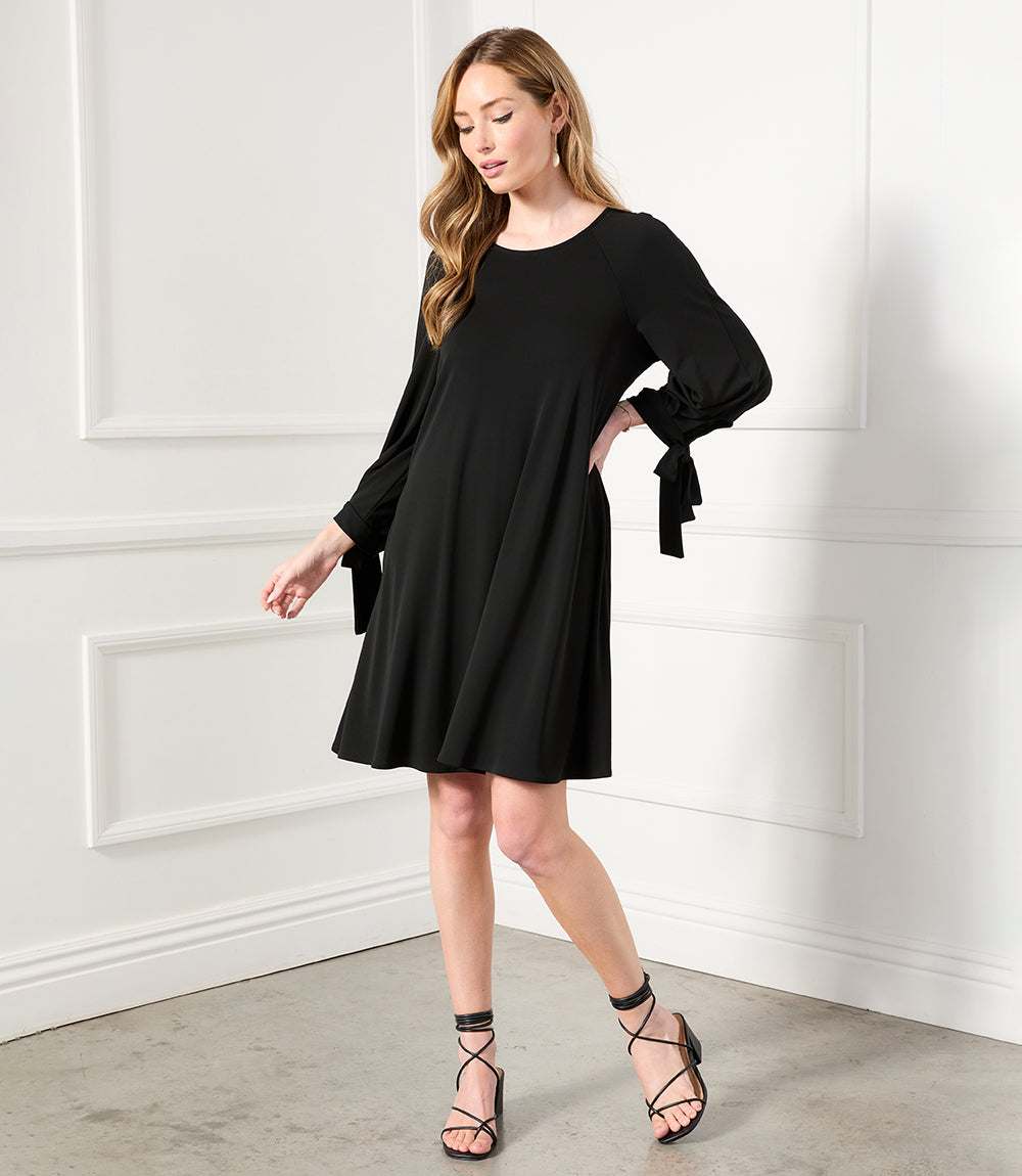 Woman wearing a black Lily Swing Dress with three-quarter raglan sleeves and tie details at cuffs.