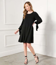 Woman wearing a black Lily Swing Dress with three-quarter raglan sleeves and tie details at cuffs.