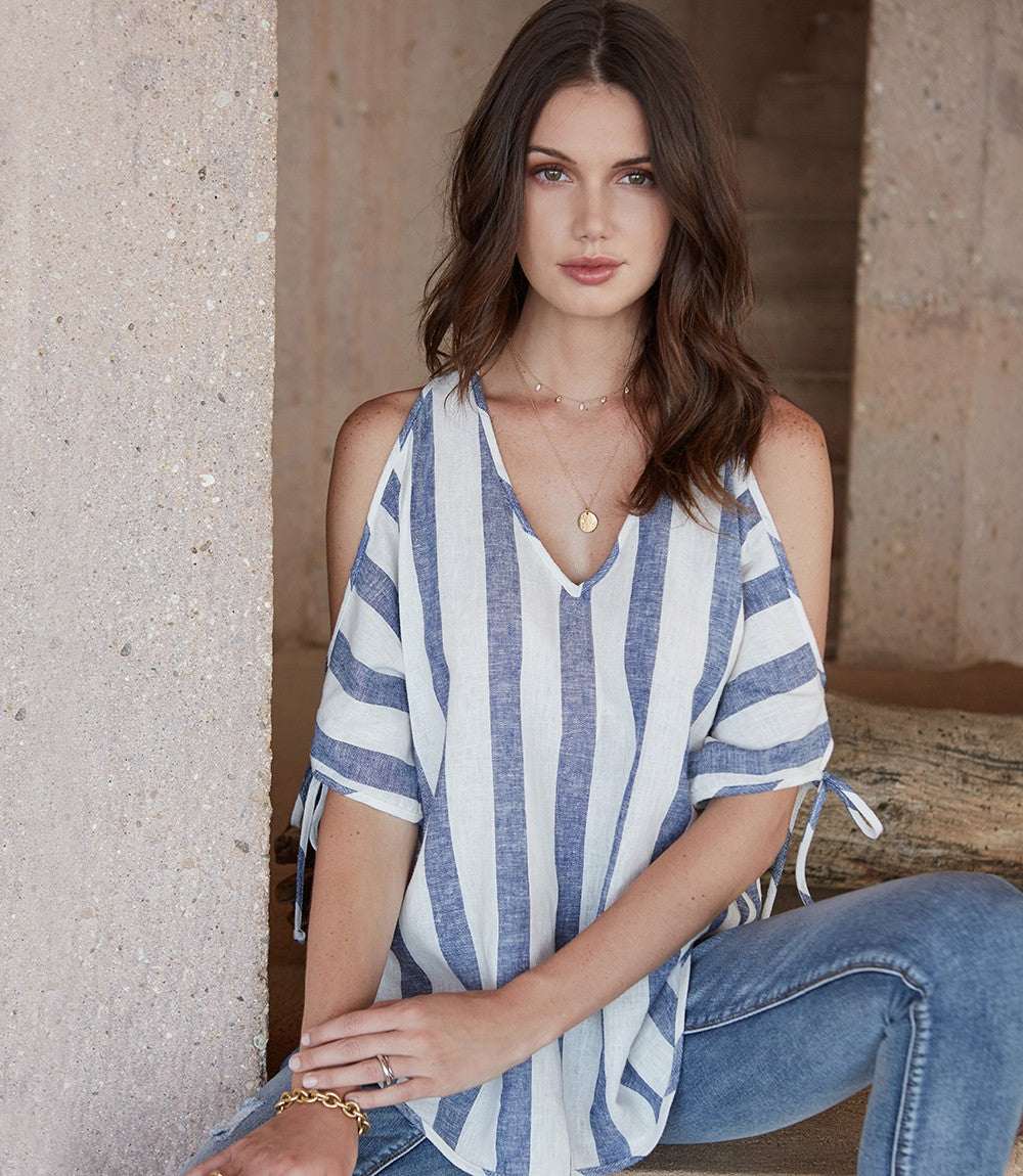 Woman wearing a blue and white striped cold shoulder tie sleeve top with a V-neck and shirttail hem.